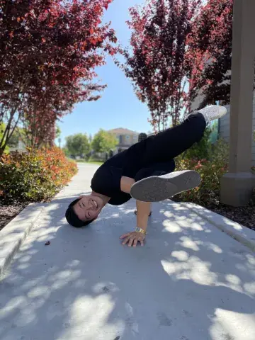 Robert Yu doing a sideways handstand on the sidewalk, red trees frame the scene from the background.