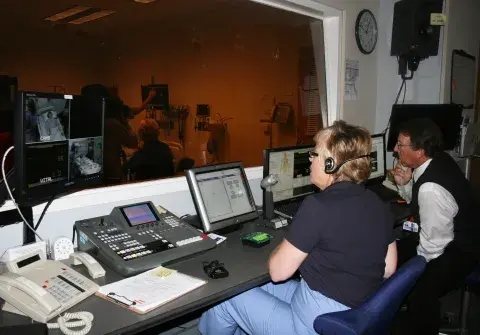 Two people in a control room looking through window at a simulation training.