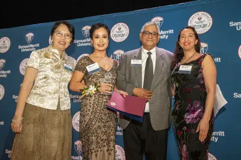 Photo of four individuals standing together. From left to right: a woman in a gold blouse and brown skirt, a woman in black, a man in a suit with a boutonnière, and a woman with white hair and glasses wearing a brown jacket. The background shows a banner for Samuel Merritt University.