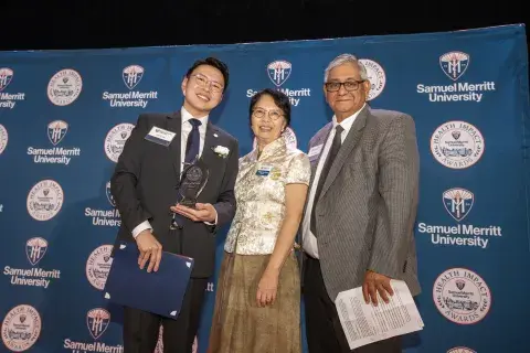 Photo of three individuals on stage at an awards ceremony. The man in the center is holding an award trophy, and the woman on the left and man on the right are standing next to him. They are smiling, and the background is a blue banner with the ‘Samuel Merritt University Health Impact Awards’ logo.