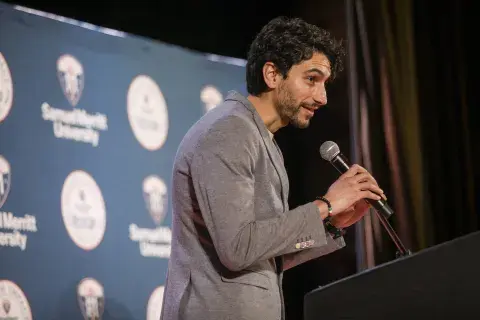Close-up of a man speaking at a podium, holding a microphone. He has curly dark hair, a light gray blazer, and is smiling while addressing the audience. The background features the ‘Samuel Merritt University’ logo on a blue banner.