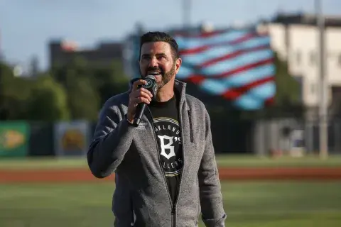 A man in an Oakland Ballers t-shirt and Samuel Merritt University jacket sings National Anthem into a microphone on the baseball field, with an American flag backdrop waving in the distance.