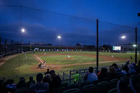 A wide view of the Oakland Ballers baseball field at dusk, with players in action and fans watching from the stands under the stadium lights.