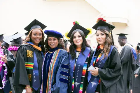 Dr. Paulina Van in academic regalia poses with three graduates in black gowns and caps, each wearing colorful stoles and cords, smiling proudly at commencement.