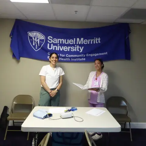 Two women stand in front of a Samuel Merritt University banner that reads “Center for Community Engagement – Ethnic Health Institute.” One woman wears a white shirt and green pants, while the other, in pink scrubs and a white cardigan, holds a clipboard. A table with health screening equipment, including a blood pressure cuff, sits in front of them.