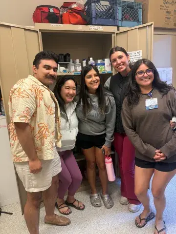 A group of five people standing close together indoors, smiling in front of open shelves stocked with medical supplies. They are casually dressed, and one person wears a Samuel Merritt University sweatshirt.
