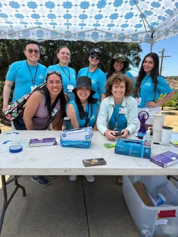 Eight people in matching blue t-shirts smiling together under a white and blue patterned canopy. They are gathered around a table with boxes of gloves, tape, and medical supplies on it, outdoors in a sunny setting.