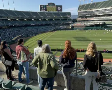 Six women stand at the railing of the Oakland Coliseum during a Soul SC match.