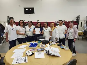 A group of eight nursing students and faculty, all in scrubs or white coats, stand together smiling behind a table covered with medical supplies, health education materials, and blood pressure cuffs. The event takes place in a gymnasium with other booths visible in the background.