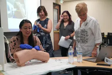 A woman wearing blue medical gloves practices a clinical procedure on a medical training mannequin in a classroom setting. She is seated at a table with two water bottles and papers, while three other women stand nearby, watching and smiling. A presentation slide is projected in the background.