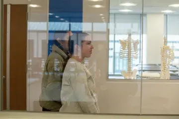Two students walk through hallway with skeleton in classroom behind window
