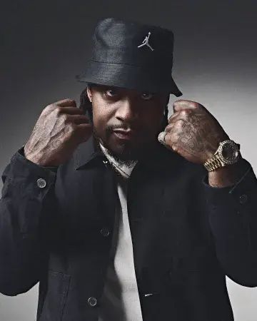 Studio portrait of a Marshawn Lynch wearing a black bucket hat with a white Jumpman logo, black jacket, and white shirt, raising both fists near his face. He has visible hand tattoos and is wearing a gold watch and ring, looking directly at the camera against a dark background.