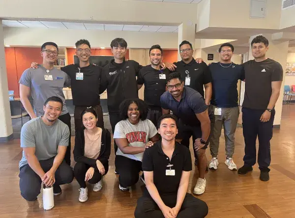 A group of twelve people, including physical therapy students and staff, pose together indoors in a bright room with wood floors and a warm-toned wall. Some are standing while others are kneeling or sitting in front, all smiling at the camera.