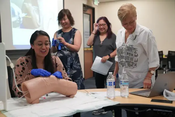 A woman wearing blue medical gloves practices a clinical procedure on a medical training mannequin in a classroom setting. She is seated at a table with two water bottles and papers, while three other women stand nearby, watching and smiling. A presentation slide is projected in the background.