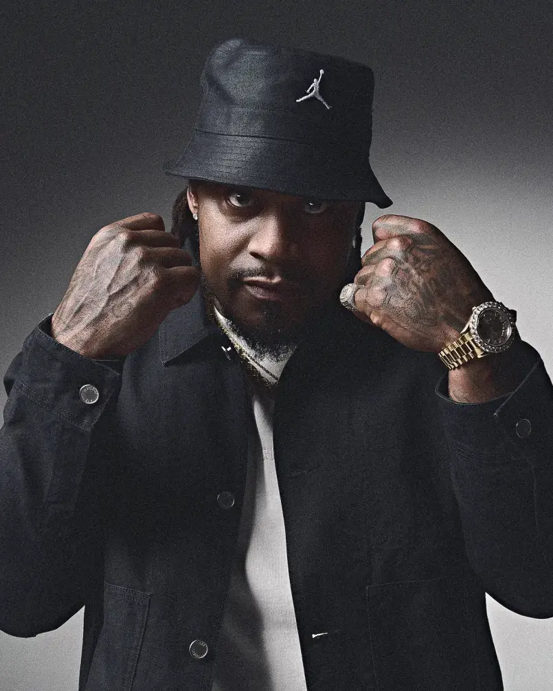 Studio portrait of a Marshawn Lynch wearing a black bucket hat with a white Jumpman logo, black jacket, and white shirt, raising both fists near his face. He has visible hand tattoos and is wearing a gold watch and ring, looking directly at the camera against a dark background.
