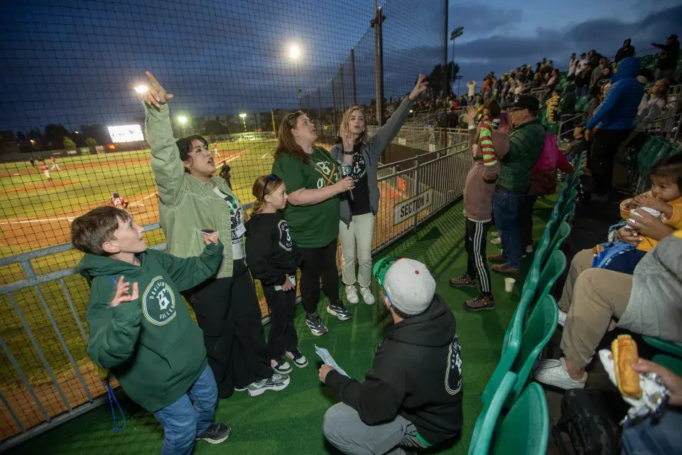 A lively group of Samuel Merritt University attendees, including children and adults in Oakland Ballers gear, cheer from the stands during the game.