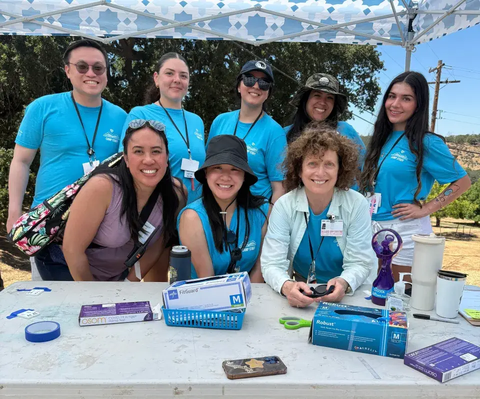 Eight people in matching blue t-shirts smiling together under a white and blue patterned canopy. They are gathered around a table with boxes of gloves, tape, and medical supplies on it, outdoors in a sunny setting.
