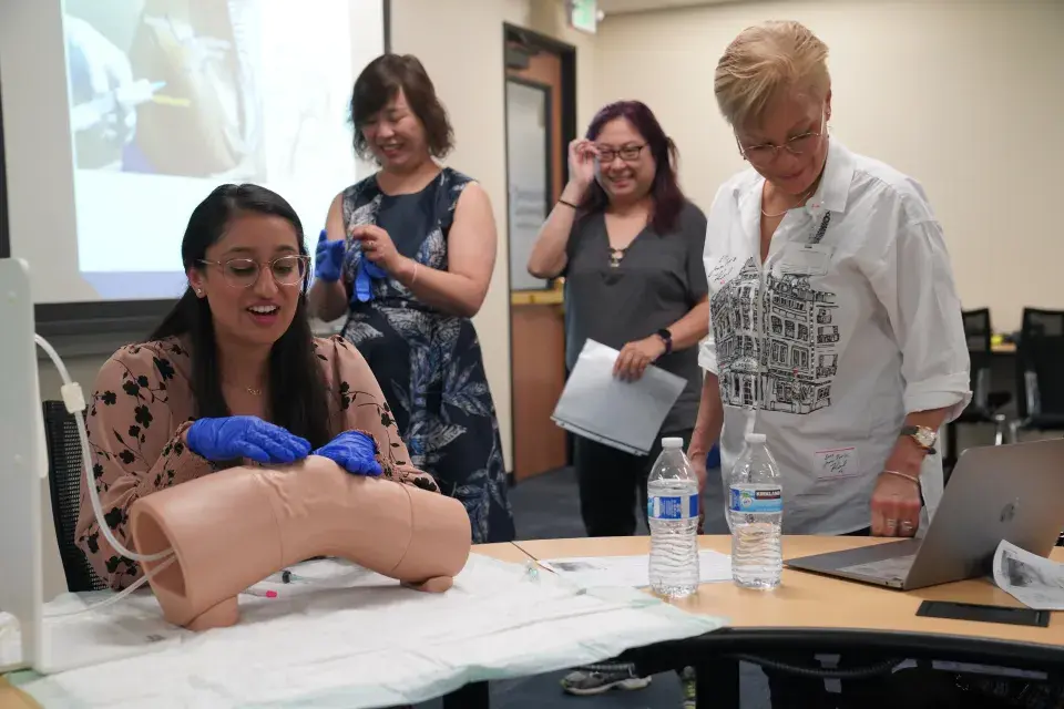 A woman wearing blue medical gloves practices a clinical procedure on a medical training mannequin in a classroom setting. She is seated at a table with two water bottles and papers, while three other women stand nearby, watching and smiling. A presentation slide is projected in the background.