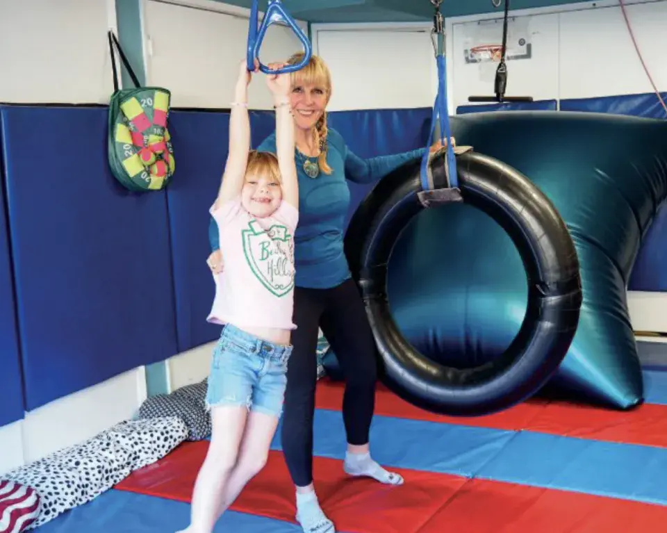 A smiling child holds onto a blue swing handle in a colorful therapy room, supported by an adult standing behind her. The adult, dressed in a teal long-sleeve shirt and black leggings, steadies the child as she hangs with both arms extended. The room is padded with blue walls and red-and-blue mats, with therapy equipment including a large inflated black ring and a soft target game visible in the background.