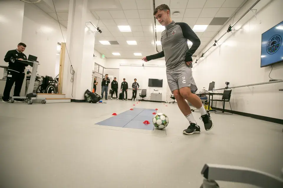 Open laboratory with athlete kicking soccer ball. Other stand in the background assessing.