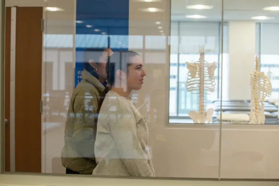 Two students walk through hallway with skeleton in classroom behind window