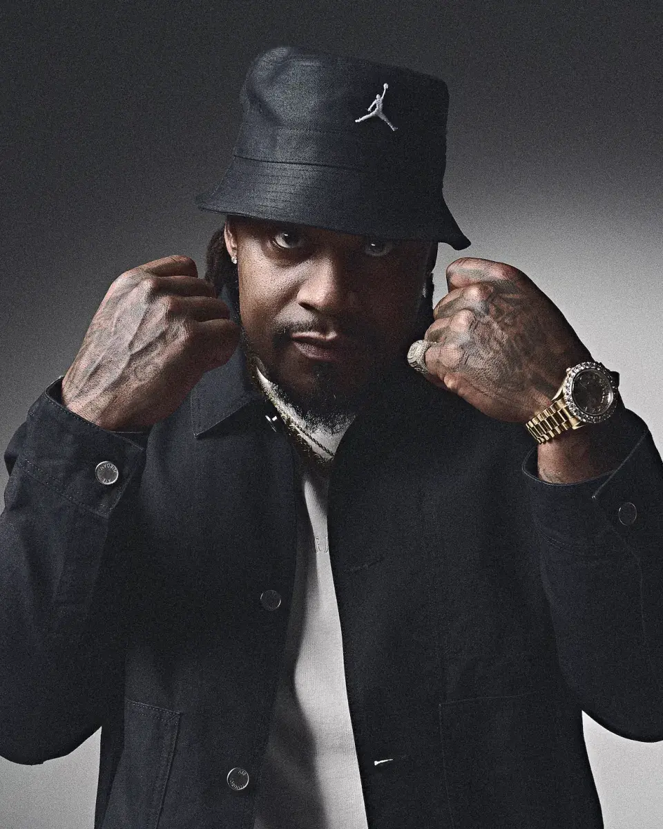 Studio portrait of a Marshawn Lynch wearing a black bucket hat with a white Jumpman logo, black jacket, and white shirt, raising both fists near his face. He has visible hand tattoos and is wearing a gold watch and ring, looking directly at the camera against a dark background.