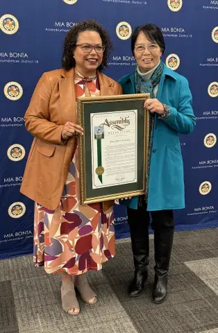 Mia Bonta and President Wang standing in front of a blue background holding framed resolution