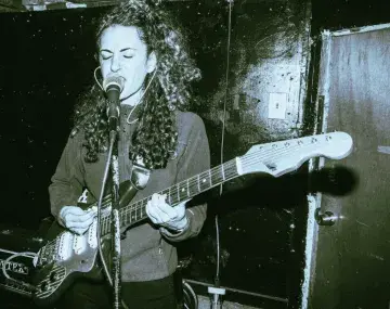 A person with long curly hair passionately sings into a microphone while playing an electric guitar in a dimly lit venue. They wear a zip-up jacket and appear deeply focused, with eyes closed mid-performance. The scene has a gritty, raw aesthetic, with a textured black wall and a visible amplifier in the background, capturing the intensity and emotion of a live alternative music performance.