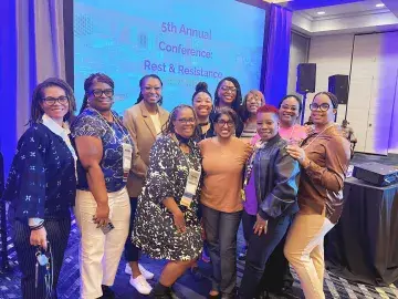 A group of 11 conference attendees smile and pose together in a meeting room. A presentation screen in the background reads “5th Annual Conference: Rest & Resistance.”