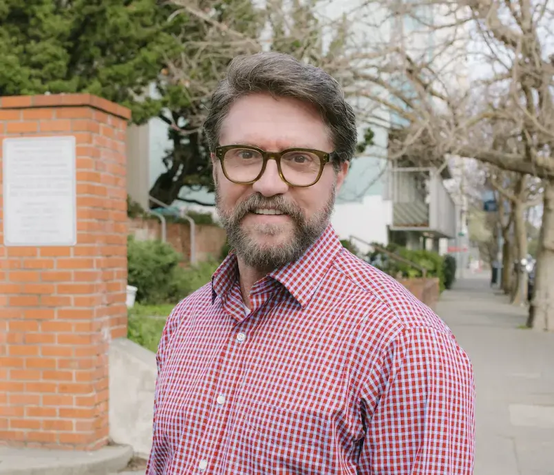 Dean Steven Rush standing outdoors wearing a red checkered shirt and glasses as he smiles.