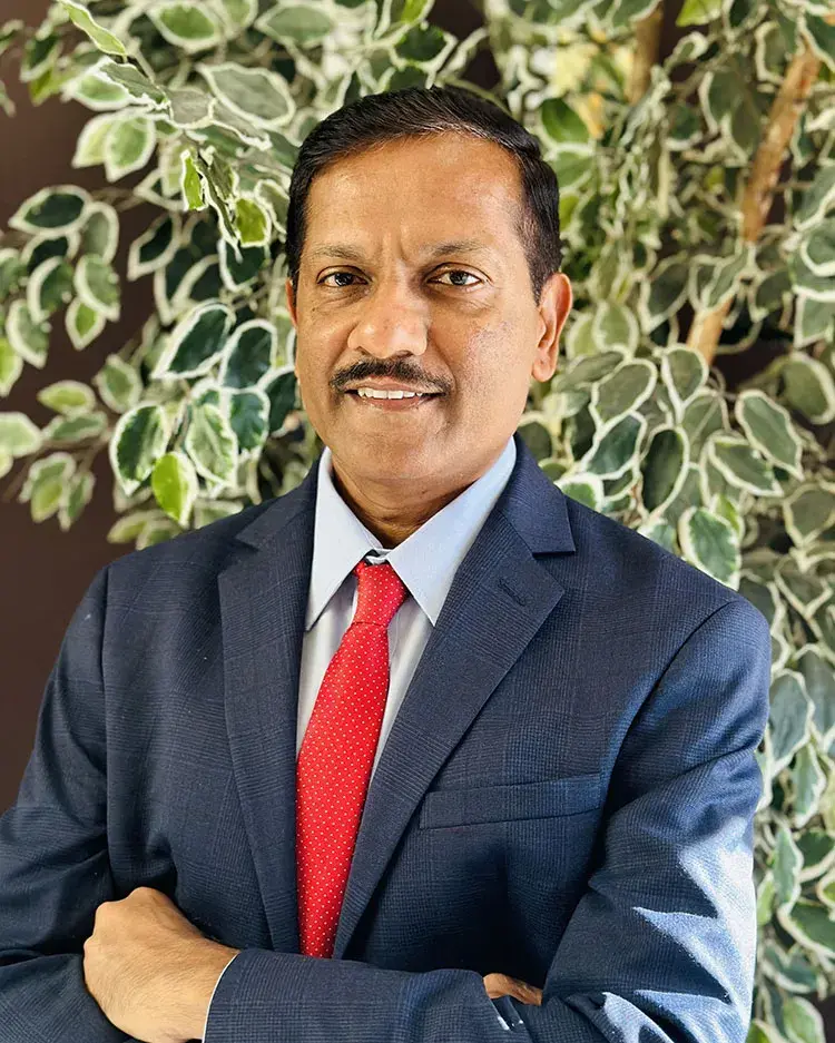 College of Health Sciences new dean Dr. Chandra stands in front of leafy background wearing blue suit and red tie.